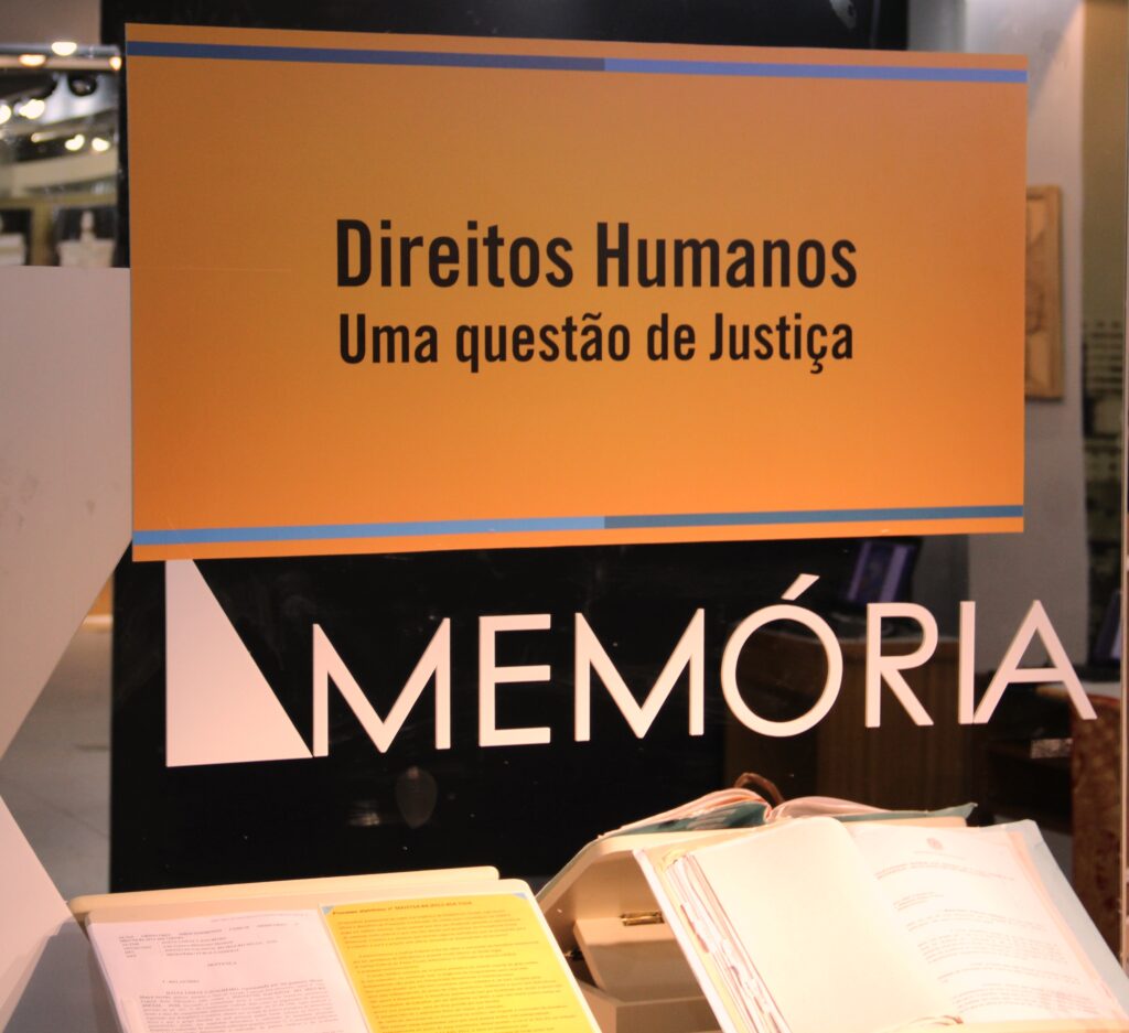 Dois processos abertos, expostos na frente de uma parte de vidro do memorial. Está escrito no adesivo colocado ao vidro: Direitos Humanos - Uma questão de justiça. MEMÓRIA.