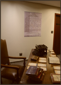 Uma cadeira e uma mesa antigas com livros raros em cima; alguns empilhados, outros abertos. E uma máquina de escrever antiga. Ao fundo, um cartaz com texto escrito .
