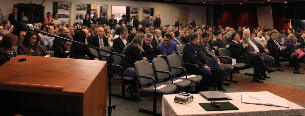 Imagem do auditório da justiça federal, praticamente lotado; há apenas com alguns lugares disponíveis mais à frente. Pessoas sentadas aguardando a palestra.
