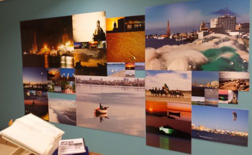 Parede com mosaico de fotos no espaço de Rio Grande - nas fotos observa-se paisagens de lugares da cidade de Rio Grande: o porto, o litoral, barcos, pôs do sol, etc.