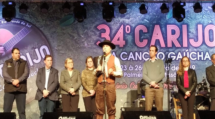 a foto mostra o palco do evento com um homem pilchado falando ao microfone. ao lado dele estão algumas pessoas, entre elas, a juíza.