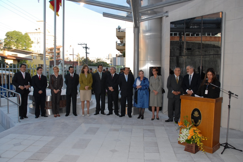 a foto mostra a solenidade de inauguração sendo realizada na frente do prédio. diversas autoridades estão posicionadas uma ao lado da outra enquanto uma mulher fala no parlatório