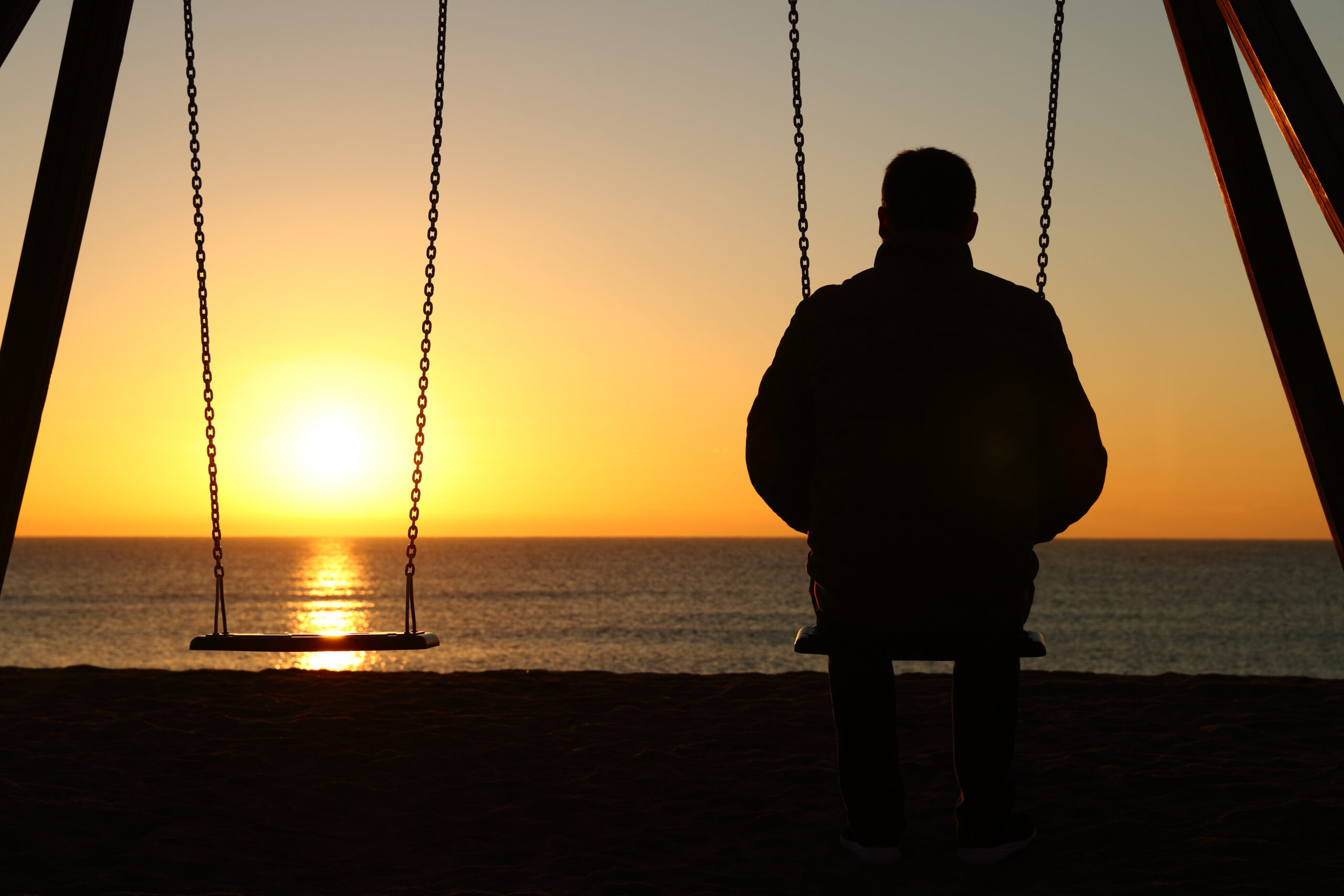 a foto mostra um homem sentando num balanço vendo o pôr do sol. do lado dele, tem um balanço sem ninguém